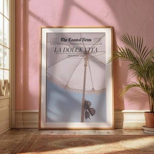 May include: A framed print of a newspaper with the headline "La Dolce Vita: The Sweet Life". The image shows a white and blue striped beach umbrella with sunglasses hanging from the handle. The background is a light blue sky.