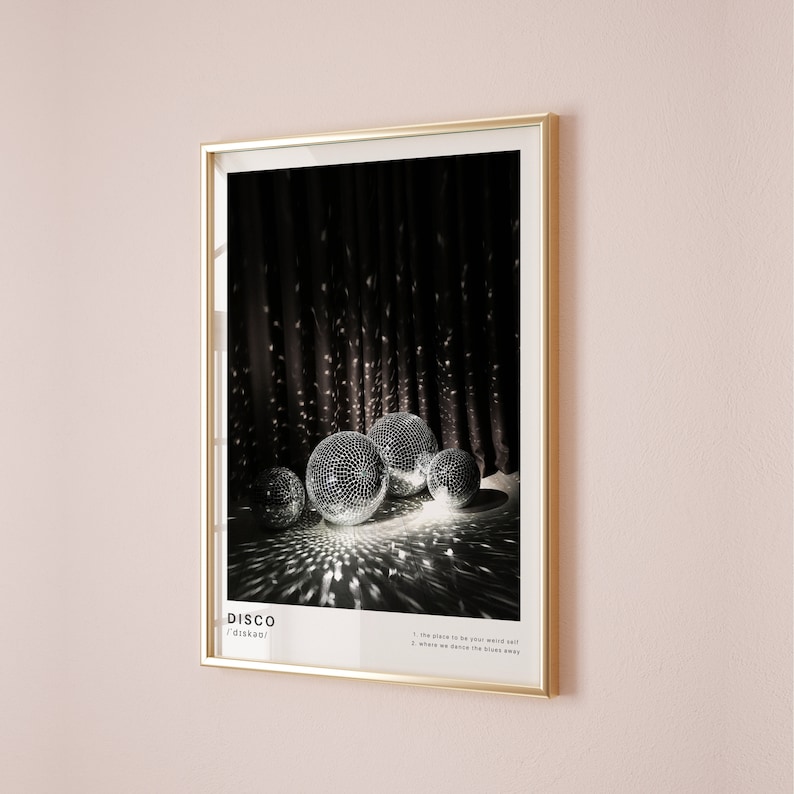 May include: A framed poster featuring a black and white photograph of disco balls against a dark backdrop. The poster displays the word "DISCO" in bold white lettering and the text "/ˈdɪskoʊ/" in smaller white lettering. The poster also includes the text "1. the place to be your weird self 2. where we dance the blues away".