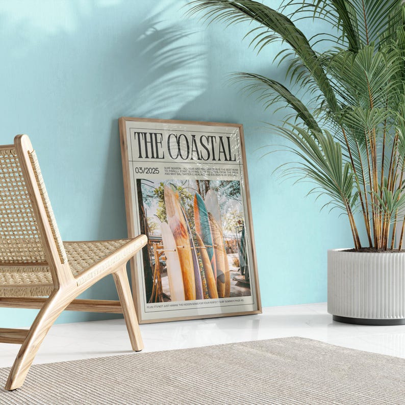 May include: A framed print titled "THE COASTAL" featuring surfboards in a beach scene. The print is in a light wood frame, leaning against a light blue wall. A woven chair and potted plant are in the foreground.