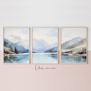 May include: A set of three watercolour paintings depicting a mountain lake scene. The paintings are framed in natural wood frames and feature a blue and grey colour palette.