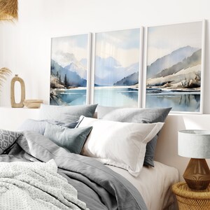 May include: A bedroom interior with a grey and white bedding set, a white lamp with a grey shade, and three framed watercolour paintings of a mountain lake scene.
