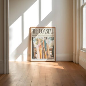 May include: A framed poster titled "THE COASTAL" featuring a photograph of several surfboards leaning against a wooden structure. The poster has a light brown wooden frame and is leaning against a white wall. The date "03/2025" is printed on the poster.