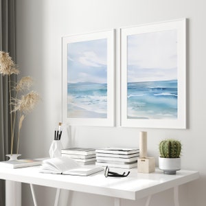 May include: Two framed watercolor paintings of a seascape with blue waves and a white sandy beach. The paintings are hanging on a white wall above a white desk with books, a vase, and a cactus.