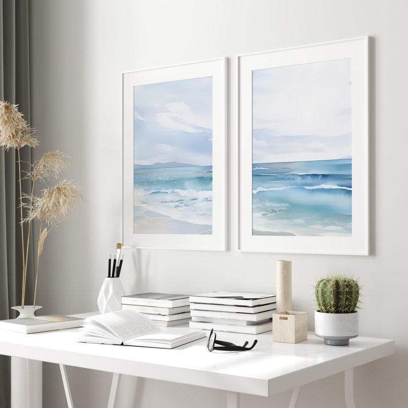 May include: Two framed watercolor paintings of a seascape with blue waves and a white sandy beach. The paintings are hanging on a white wall above a white desk with books, a vase, and a cactus.