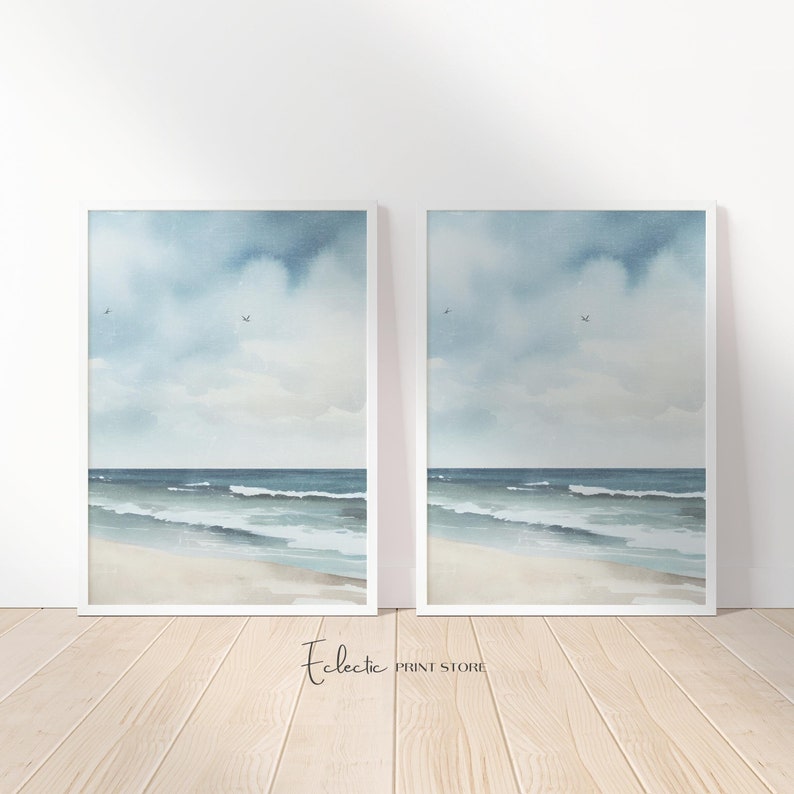 May include: Two framed watercolor paintings of a seascape with a sandy beach, blue ocean, and a cloudy sky. The paintings are in white frames and are hanging on a white wall. The text 'Eclectic Print Store' is visible at the bottom of the image.