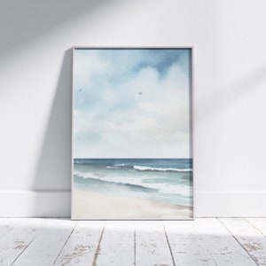 May include: A framed watercolor painting of a seascape. The painting depicts a blue sky with white clouds, a blue ocean with white waves, and a sandy beach. The painting is framed in a light gray frame.