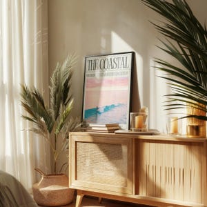 May include: A framed poster with the text "THE COASTAL" and a coastal scene, resting on a light wood cabinet. The cabinet has a woven panel and is decorated with candles and books. A potted plant and a basket are also in the image.