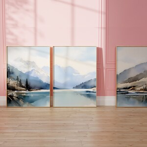 May include: Three framed watercolour paintings of a mountain lake scene. Each painting features a blue lake with mountains in the background. The paintings are framed in gold.