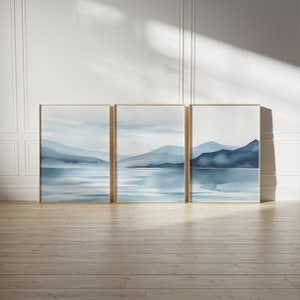 Könnte beinhalten: Drei gerahmte Aquarellbilder, die eine blaue und weiße Landschaft mit Bergen und Wasser darstellen. Die Bilder sind in goldenen Rahmen gefasst und an einer weißen Wand aufgehängt.
