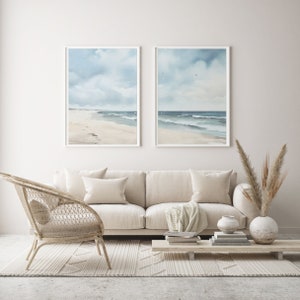 May include: Two framed watercolor paintings of a beach scene with blue skies and white clouds. The paintings are hanging on a white wall above a white couch with two pillows and a throw blanket. A wicker chair and a wooden coffee table with books and a vase of dried flowers are in front of the couch. The floor is covered in a white rug with a geometric pattern.