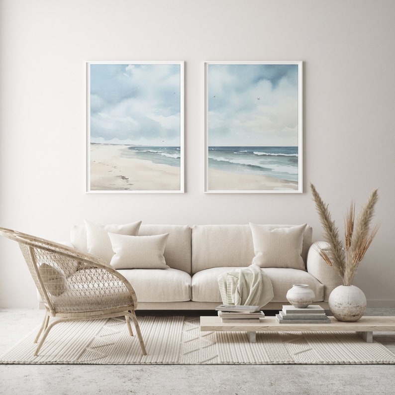 May include: Two framed watercolor paintings of a beach scene with blue skies and white clouds. The paintings are hanging on a white wall above a white couch with two pillows and a throw blanket. A wicker chair and a wooden coffee table with books and a vase of dried flowers are in front of the couch. The floor is covered in a white rug with a geometric pattern.