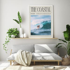 May include: A framed print titled "THE COASTAL" featuring a surfer riding a large wave. The image has a light blue and white color scheme. The print is displayed above a gray sofa with a wooden frame, with plants and decor.