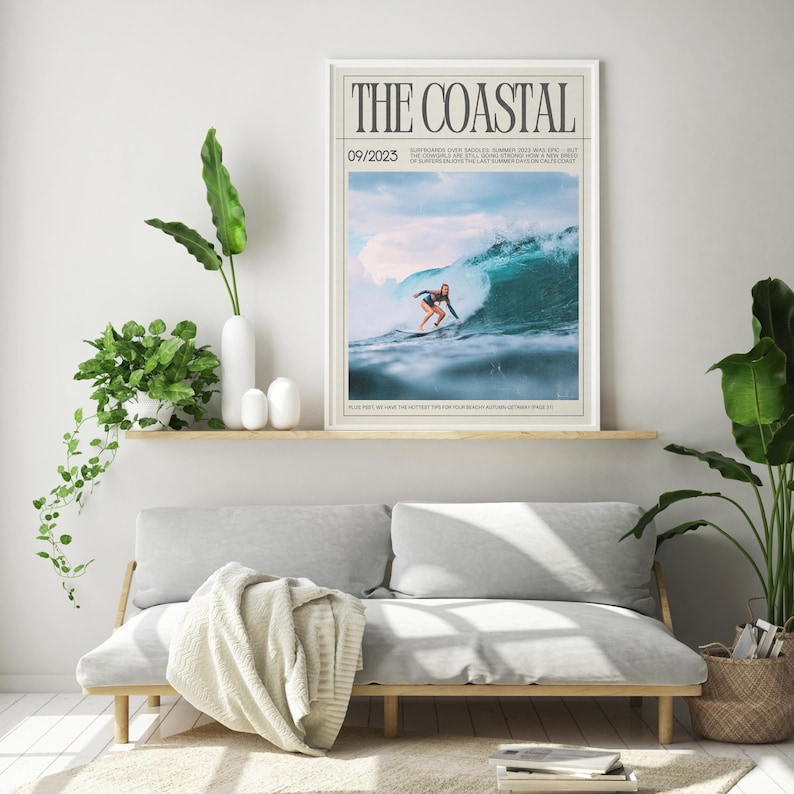 May include: A framed print titled "THE COASTAL" featuring a surfer riding a large wave. The image has a light blue and white color scheme. The print is displayed above a gray sofa with a wooden frame, with plants and decor.