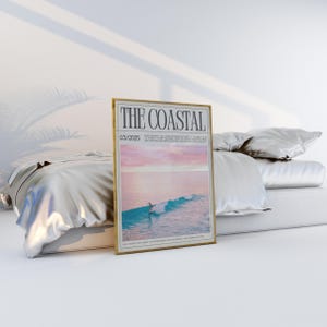 May include: A framed poster titled "THE COASTAL" with a gold frame. The poster features a surfer on a wave with a pink and blue sky. The poster is displayed on a bed with white bedding.