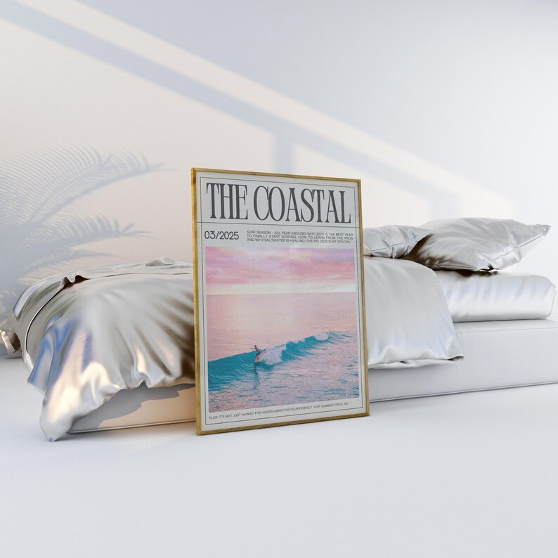 May include: A framed poster titled "THE COASTAL" with a gold frame. The poster features a surfer on a wave with a pink and blue sky. The poster is displayed on a bed with white bedding.