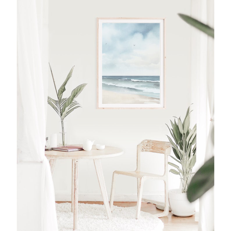 May include: A framed watercolor painting of a beach scene with blue water, white sand, and a light blue sky with clouds. The painting is hanging on a white wall in a room with a white table and chair.