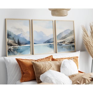 May include: Three framed watercolour paintings depicting a mountain lake scene with blue and grey mountains, a blue lake, and a light blue sky. The paintings are hung on a white wall above a bed with brown and white pillows.