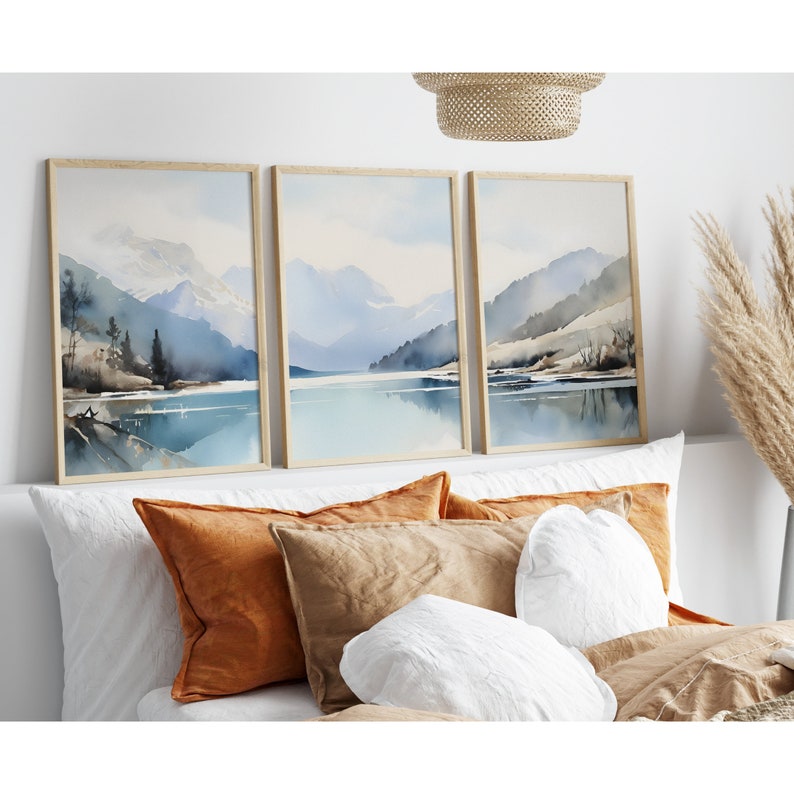 May include: Three framed watercolour paintings depicting a mountain lake scene with blue and grey mountains, a blue lake, and a light blue sky. The paintings are hung on a white wall above a bed with brown and white pillows.