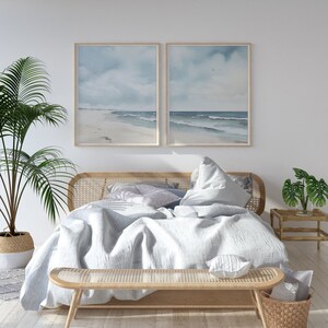May include: A light wood bed frame with a white linen duvet and pillows. The bed is decorated with a light blue and white throw pillow and a woven bench at the foot of the bed. Two framed watercolor paintings of a beach scene are hanging on the wall behind the bed.