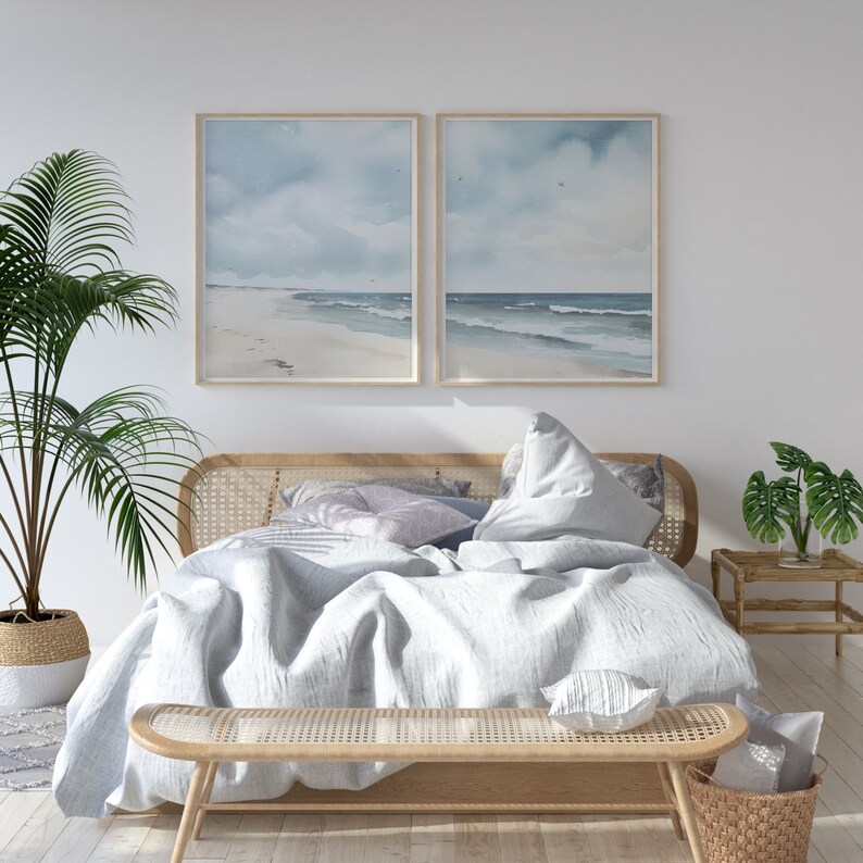May include: A light wood bed frame with a white linen duvet and pillows. The bed is decorated with a light blue and white throw pillow and a woven bench at the foot of the bed. Two framed watercolor paintings of a beach scene are hanging on the wall behind the bed.