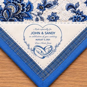May include: A quilted fabric corner featuring a blue and white floral pattern with a triangular cream-colored patch. The patch displays a blue heart graphic, two interlocking rings, and the text: Made especially for JOHN & SANDY in celebration of your wedding AUGUST 3, 2021 Love Aunt Jane.