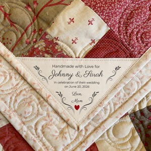 Peut inclure: Une couverture en tissu matelassé avec un motif patchwork dans des tons rouge, crème et beige, ornée de surpiqûres. Une étiquette triangulaire est cousue, portant l'inscription : Handmade with Love for Johnny & Sarah, In celebration of their wedding on June 20, 2026, Love, Mom, avec un petit cœur rouge.
