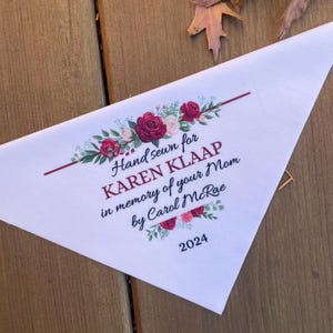 May include: A white triangular fabric piece featuring a floral design with red and pink roses and green leaves. The text reads: Hand sewn for KAREN KLAAP in memory of your Mom by Carol McRae 2024. The design includes a horizontal maroon line.