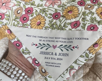 Könnte beinhalten: Ein gestepptes Stoffstück mit einem Blumenmuster in Gelb, Rosa und Grün. Ein dreieckiger Aufnäher ist aufgenäht, bedruckt mit: MAY THE THREADS THAT KEEP THIS QUILT TOGETHER BE AS STRONG AS YOUR LOVE, JESSICA & JOHN, JULY 5TH, 2024, MADE WITH LOVE SOLANGE JAMES.