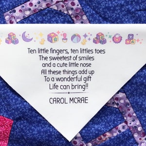 May include: A white triangular fabric patch with a poem about a baby, set on a blue and purple quilt. The poem includes the words "Ten little fingers, ten little toes" and is signed "CAROL MCRAE."