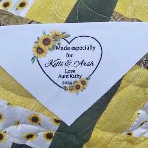 May include: White fabric label with a heart-shaped design featuring sunflowers and the text "Made especially for Kelli & Arsh Love Aunt Kathy 2024".