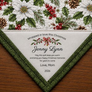 May include: A quilted fabric corner featuring a white background with a pattern of green pine branches, red berries, white poinsettias, and pinecones. A triangular label reads: Wrapped in love this Christmas, Jenny Lynn, 2026, Love, Mom.
