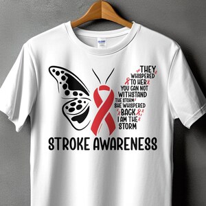 May include: White t-shirt with a red ribbon butterfly design and the text "THEY WHISPERED TO HER YOU CAN NOT WITHSTAND THE STORM SHE WHISPERED BACK I AM THE STORM STROKE AWARENESS".