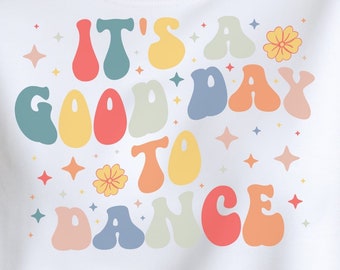 May include: A white sweatshirt with the text "It's a good day to dance" in colorful, retro-style lettering. The text is surrounded by small stars and flowers.