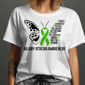 Op de afbeelding: Wit T-shirt met een zwart vlinderontwerp en een groen lint, ter bevordering van Biliaire Atresie Awareness. Het shirt heeft de tekst "THEY WHISPERED TO HER...I AM THE STORM" in zwart en groen.