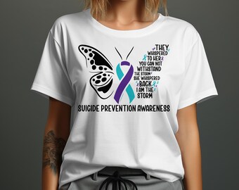 May include: White t-shirt with a butterfly design and the words "SUICIDE PREVENTION AWARENESS". The butterfly has a black wing pattern, and a purple and teal ribbon. Text on the shirt reads "THEY WHISPERED TO HER...I AM THE STORM."