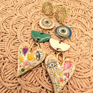 Peut inclure: Une paire de boucles d'oreilles pendantes avec des clous circulaires texturés dorés, des breloques œil porte-bonheur avec strass, des accents demi-circulaires bleu sarcelle et blancs, et des pendentifs en forme de cœur ornés de motifs peints.
