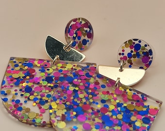 May include: A pair of dangle earrings featuring clear acrylic shapes filled with multicolored confetti dots in shades of pink, blue, and gold. Each earring consists of a small circular stud connected to a larger semi-circular pendant by a gold-tone metal piece.