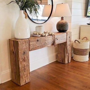 May include: A rustic wooden console table with a distressed finish, topped with a white vase holding greenery, a round mirror, and a lamp. A woven basket sits on the floor. The room has a neutral color palette.