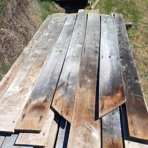 May include: A collection of weathered, reclaimed wooden planks arranged in a stack. The boards feature a rough, textured surface with visible wood grain, knots, and a mix of gray, brown, and dark charred tones from natural aging.