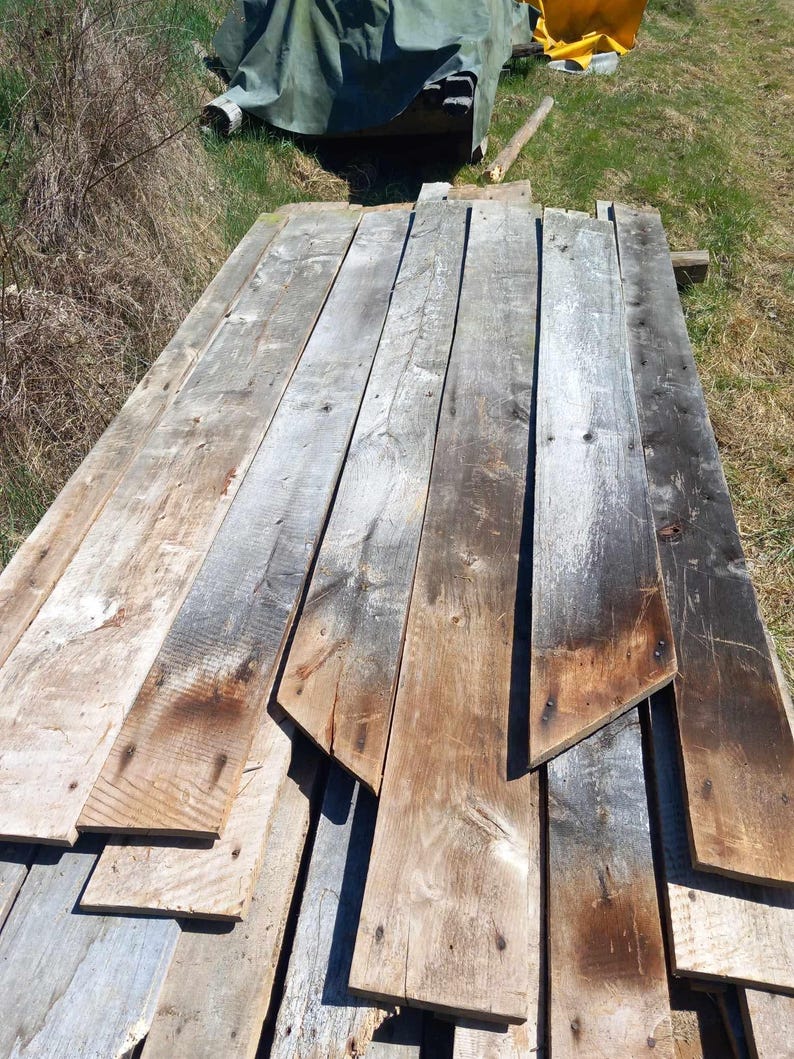 May include: A collection of weathered, reclaimed wooden planks arranged in a stack. The boards feature a rough, textured surface with visible wood grain, knots, and a mix of gray, brown, and dark charred tones from natural aging.