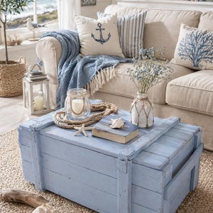 Könnte beinhalten: Ein hellblaues Holzkiste dient als Couchtisch in einem Wohnzimmer im Küstenstil. Die Truhe ist mit nautischen Dekorationen wie Seil, Seestern und einem kleinen Buch geschmückt. Kissen mit Anker- und Korallenmotiven liegen auf einem beigen Sofa und ergänzen das Meeresthema.