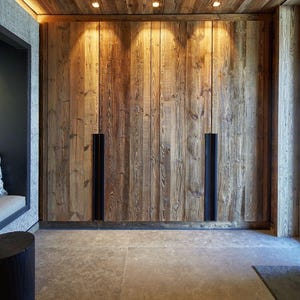 May include: A floor-to-ceiling wooden wardrobe featuring vertical planks with a natural grain texture. The unit includes two long, recessed black metal handles integrated into the doors, illuminated by three small overhead recessed lights.
