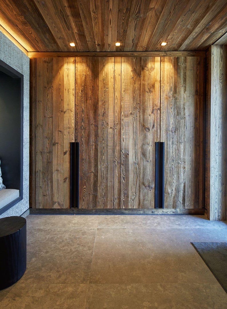 May include: A floor-to-ceiling wooden wardrobe featuring vertical planks with a natural grain texture. The unit includes two long, recessed black metal handles integrated into the doors, illuminated by three small overhead recessed lights.