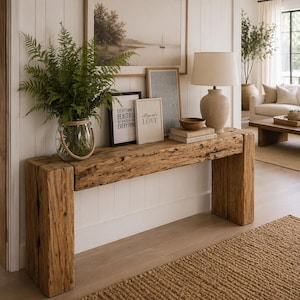 May include: A reclaimed wooden console table with a landscape painting and framed art prints. A glass vase with ferns sits on the table. A beige lamp and books are also on the table. A woven rug is on the floor.