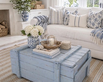 Pode incluir: Um baú de madeira azul claro serve como mesa de centro numa sala de estar. O baú é retangular com grão de madeira visível. Uma bandeja tecida com uma vela e livros está em cima, ao lado de um sofá branco com almofadas azuis.