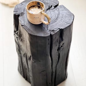 May include: A solid black wooden log stool with a natural tree trunk texture and visible grain, featuring a deep vertical crack. A speckled beige ceramic mug filled with a light-colored beverage sits on the flat top surface of the stool.