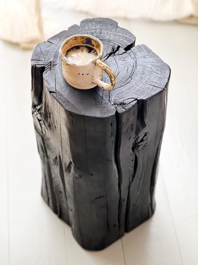 May include: A solid black wooden log stool with a natural tree trunk texture and visible grain, featuring a deep vertical crack. A speckled beige ceramic mug filled with a light-colored beverage sits on the flat top surface of the stool.