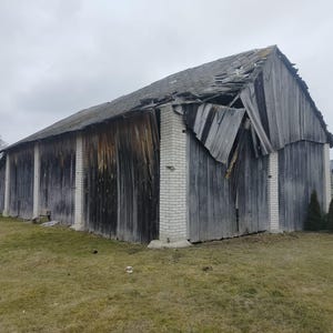 May include: A large, weathered wooden barn with vertical gray planks and white brick pillars. The roof is partially collapsed, showing damaged wooden shingles. The structure is set against a cloudy sky on a grassy field.