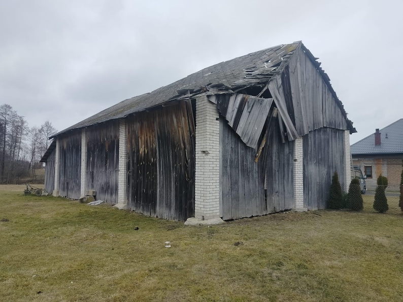 May include: A large, weathered wooden barn with vertical gray planks and white brick pillars. The roof is partially collapsed, showing damaged wooden shingles. The structure is set against a cloudy sky on a grassy field.