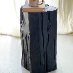 May include: A tall, cylindrical black wooden stool with a textured, natural grain finish. Resting on the flat top surface is a rectangular book with a tan cover and a speckled, light-colored ceramic mug with a handle and a dark rim.